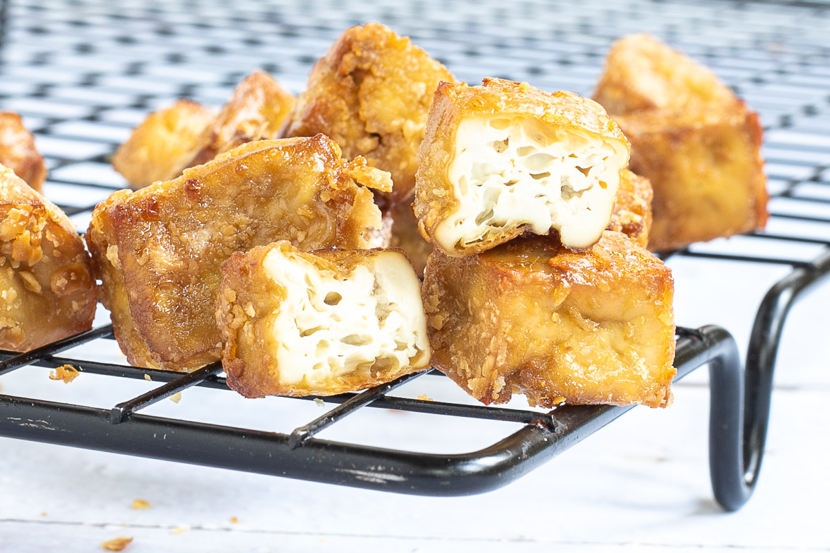 Crispy golden brown tofu cubes stacked on a black wire rack. 2 of them is cut in half to see the spongy inside. 