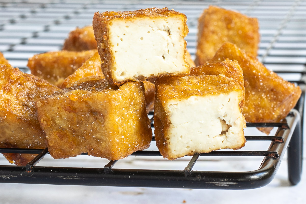 Crispy golden brown tofu cubes stacked on a black wire rack. 2 of them is cut in half to see the spongy inside.