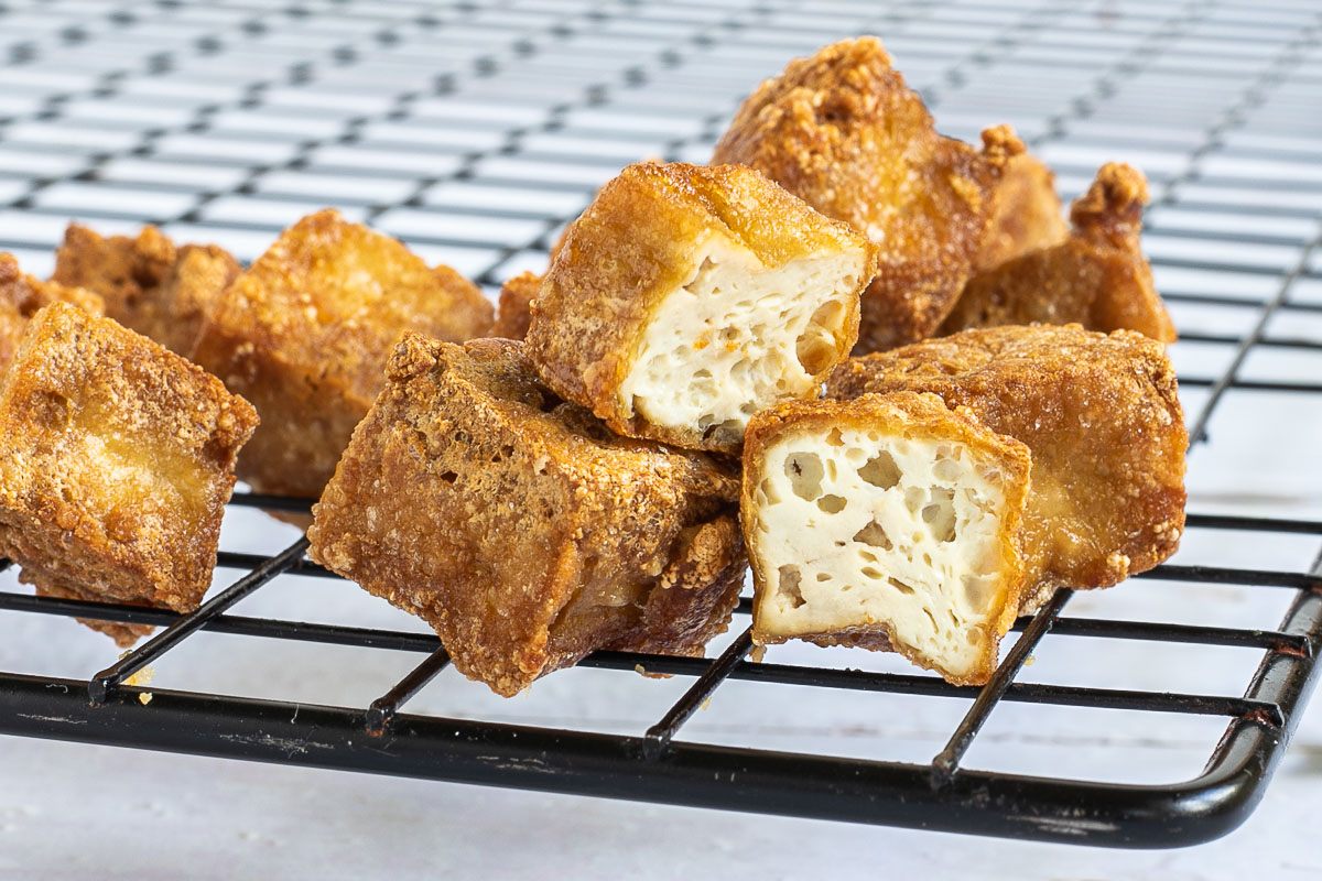 Crispy golden brown tofu cubes stacked on a black wire rack. 2 of them is cut in half to see the spongy inside.