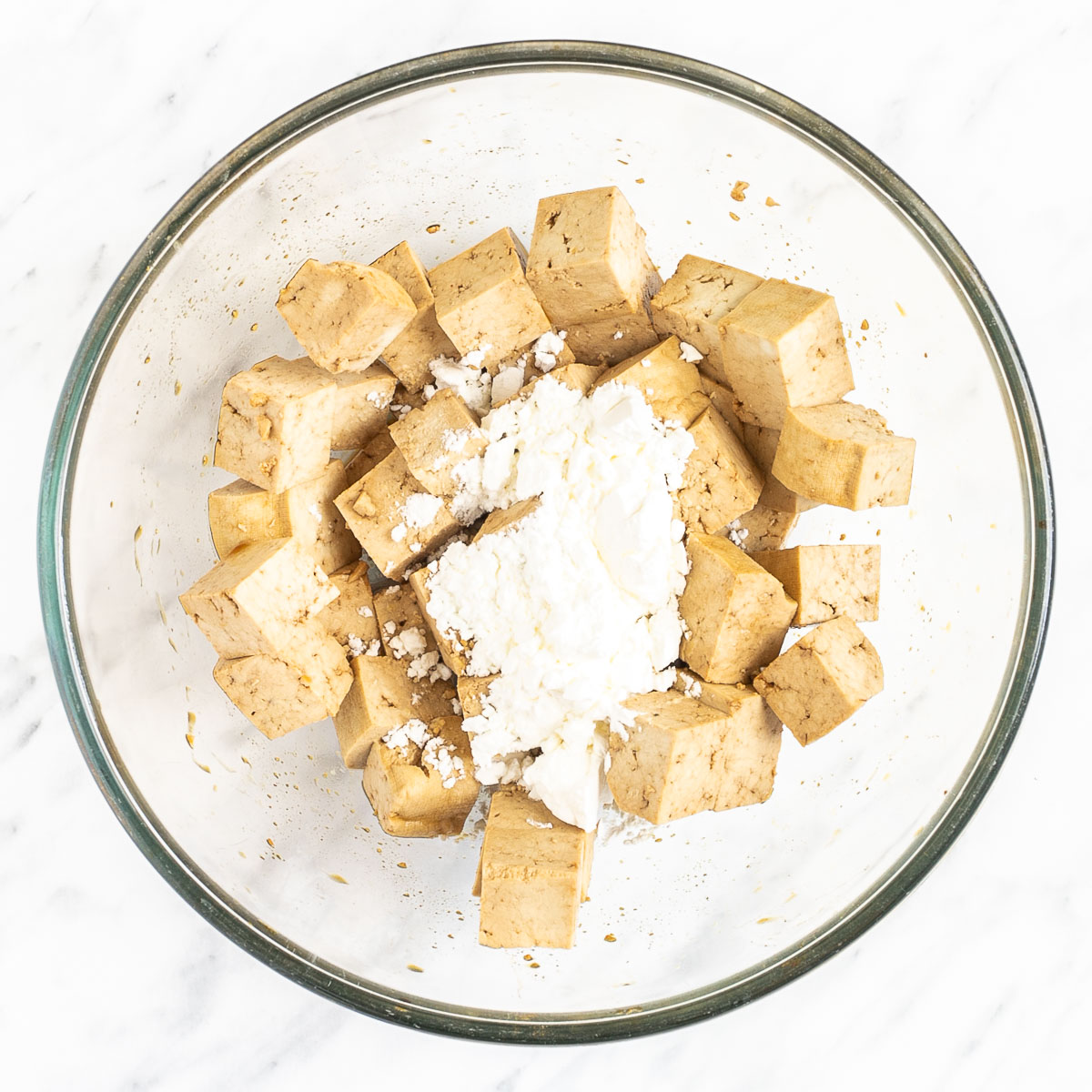 Light brown tofu cubes in a glass bowl with a heap of white powder.
