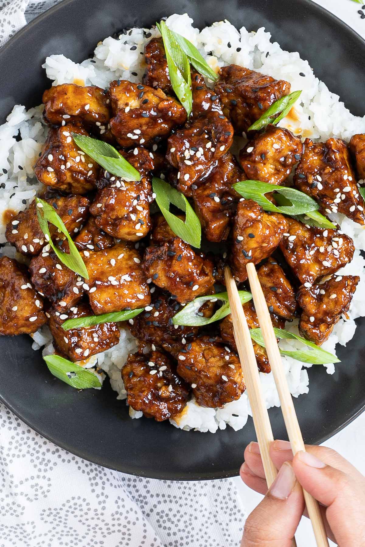 Brown sticky tofu pieces served on top of white rice on a black plate. It is sprinkled with black and white sesame seeds and scallion. A hand is holding chopsticks and about to take one.