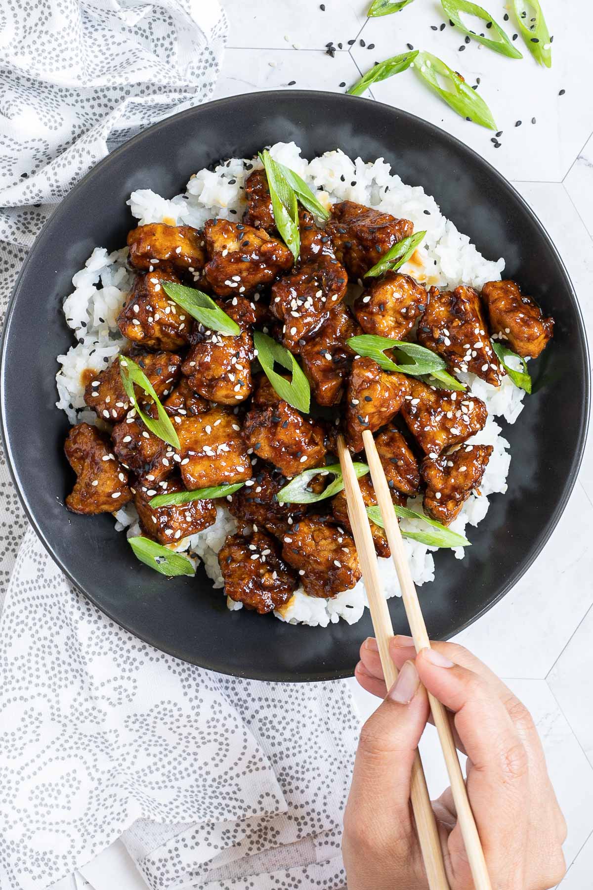 Brown sticky tofu pieces served on top of white rice on a black plate. It is sprinkled with black and white sesame seeds and scallion. A hand is holding chopsticks and about to take one. 