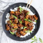 Brown sticky tofu pieces served on top of white rice on a black plate. It is sprinkled with black and white sesame seeds and scallion.