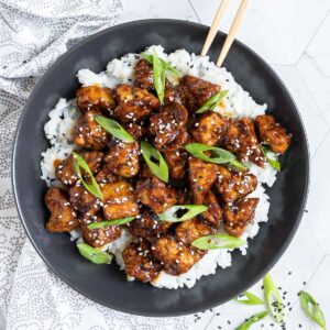 Brown sticky tofu pieces served on top of white rice on a black plate. It is sprinkled with black and white sesame seeds and scallion.
