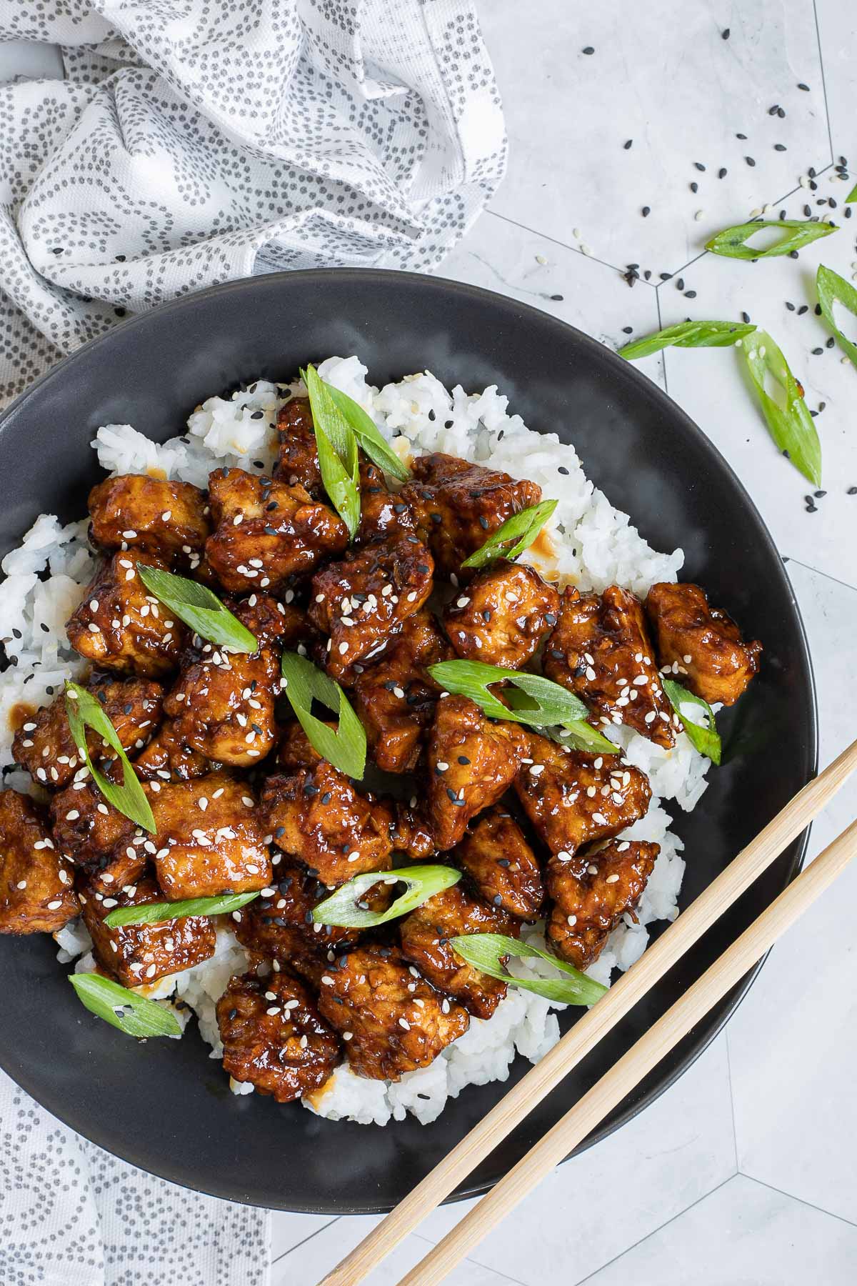 Brown sticky tofu pieces served on top of white rice on a black plate. It is sprinkled with black and white sesame seeds and scallion.