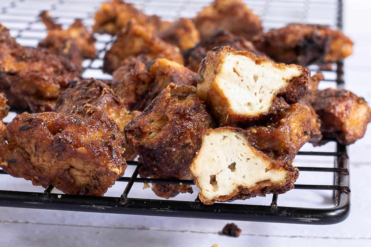 Crispy dark brown tofu cubes stacked on a black wire rack. 2 of them is cut in half to see the spongy inside.