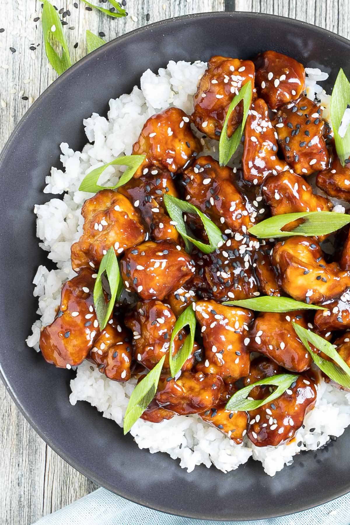Brown sticky tofu pieces served on top of white rice on a black plate. It is sprinkled with black and white sesame seeds and scallion.