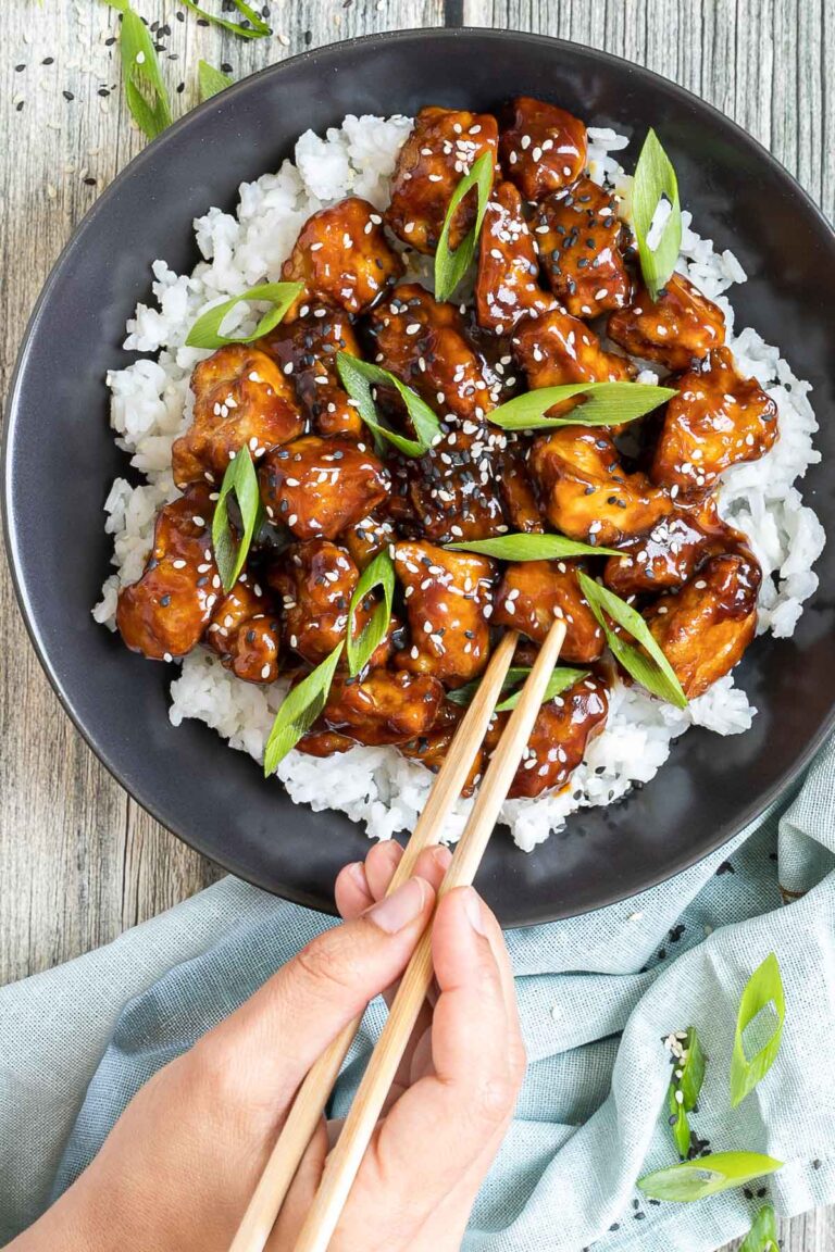 Brown sticky tofu pieces served on top of white rice on a black plate. It is sprinkled with black and white sesame seeds and scallion. A hand is holding chopsticks and is about to grab a piece.