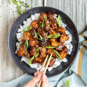 Brown sticky tofu pieces served on top of white rice on a black plate. It is sprinkled with black and white sesame seeds and scallion. A hand is holding chopsticks and is about to grab a piece.