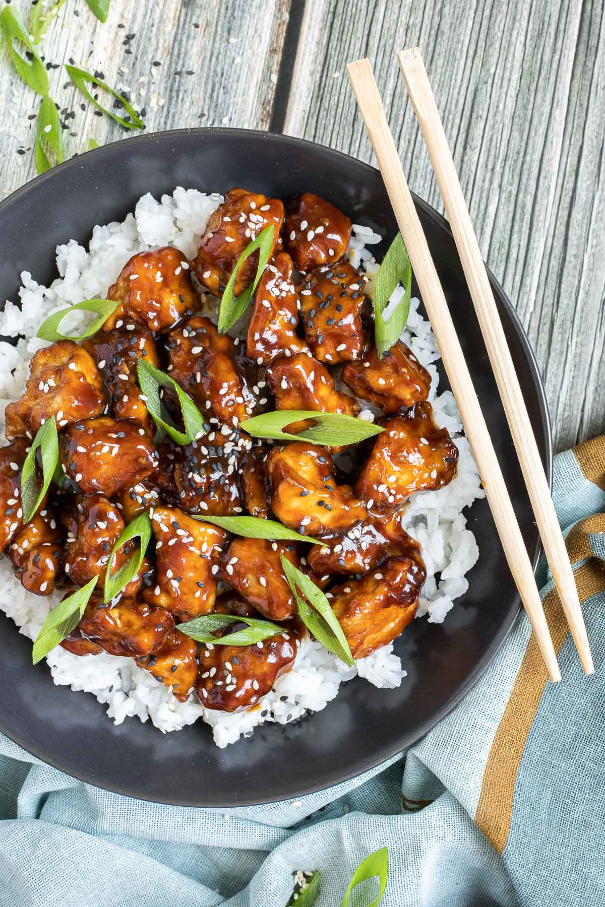 Brown sticky tofu pieces served on top of white rice on a black plate. It is sprinkled with black and white sesame seeds and scallion.
