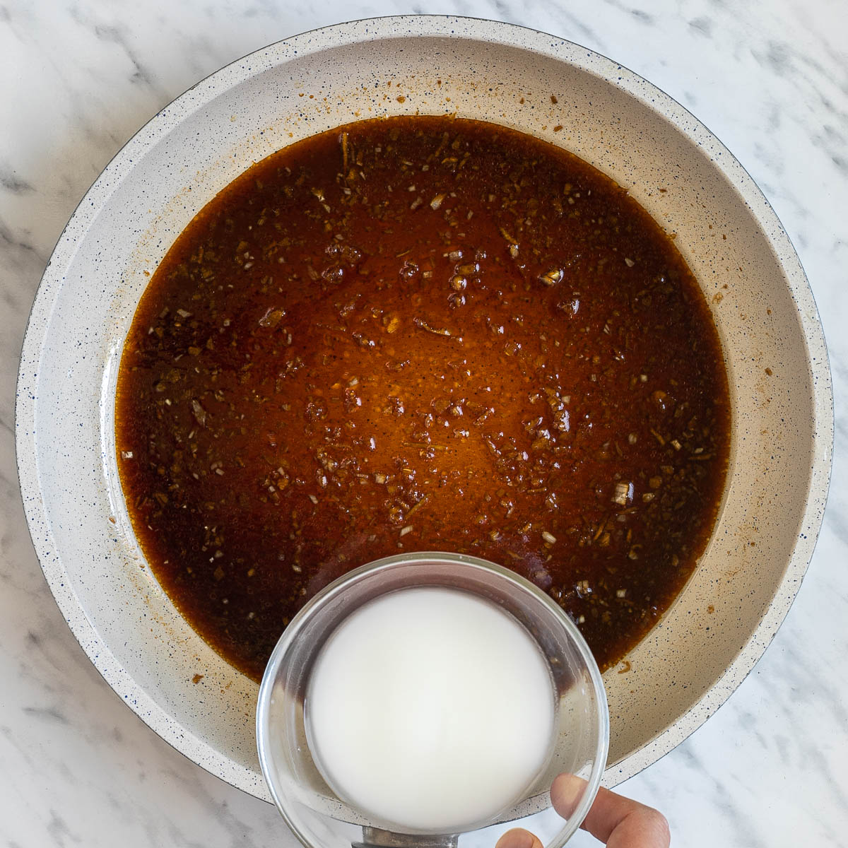 A white frying pan with different dark liquids and spices. A small bowl with white slurry is next to it.