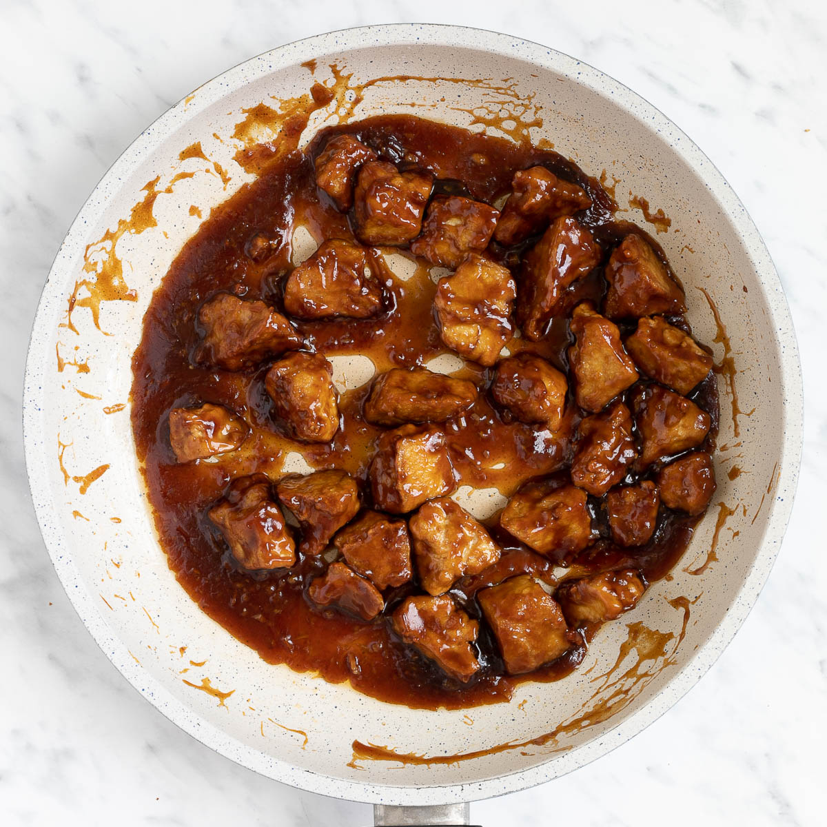 Brown sticky tofu pieces in a white frying pan.