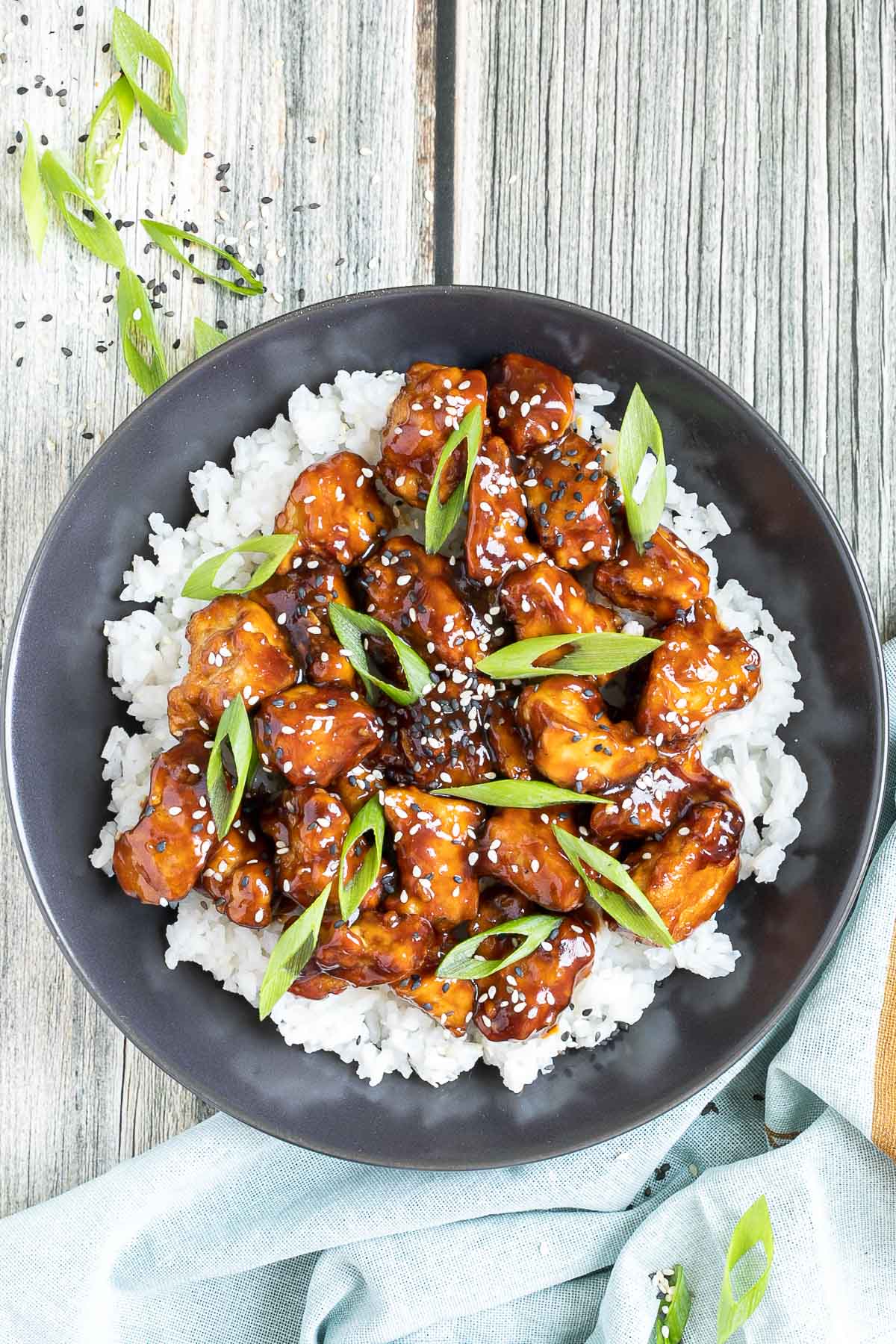 Brown sticky tofu pieces served on top of white rice on a black plate. It is sprinkled with black and white sesame seeds and scallion.