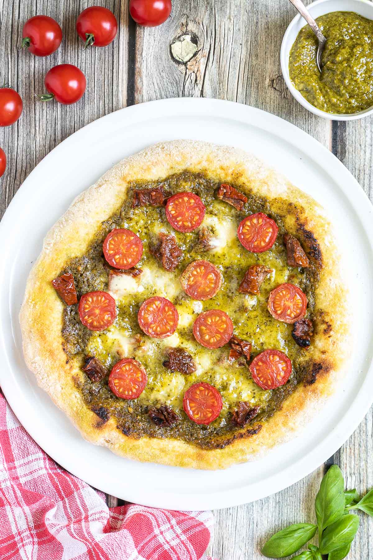 Pizza on a white pizza plate topped with green pesto, melted cheese, sun-dried tomato pieces, and halved cherry tomatoes. More pesto and fresh ingredients next to the plate.