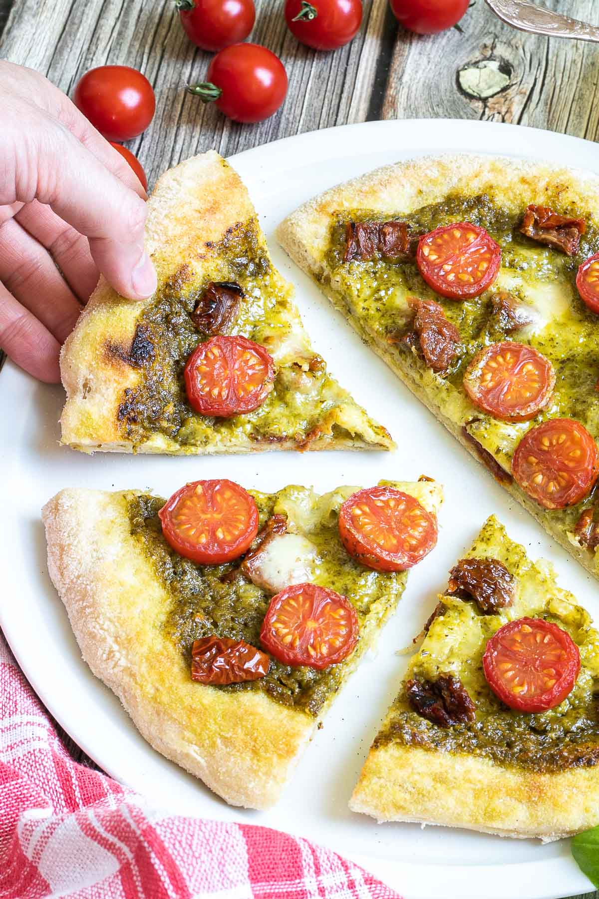 Sliced pizza on a white pizza plate topped with green pesto, melted cheese, sun-dried tomato pieces, and halved cherry tomatoes. A hand is holding one slice.