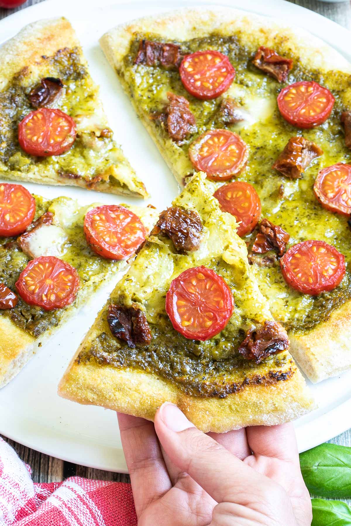 Sliced pizza on a white pizza plate topped with green pesto, melted cheese, sun-dried tomato pieces, and halved cherry tomatoes. A hand is holding one slice.