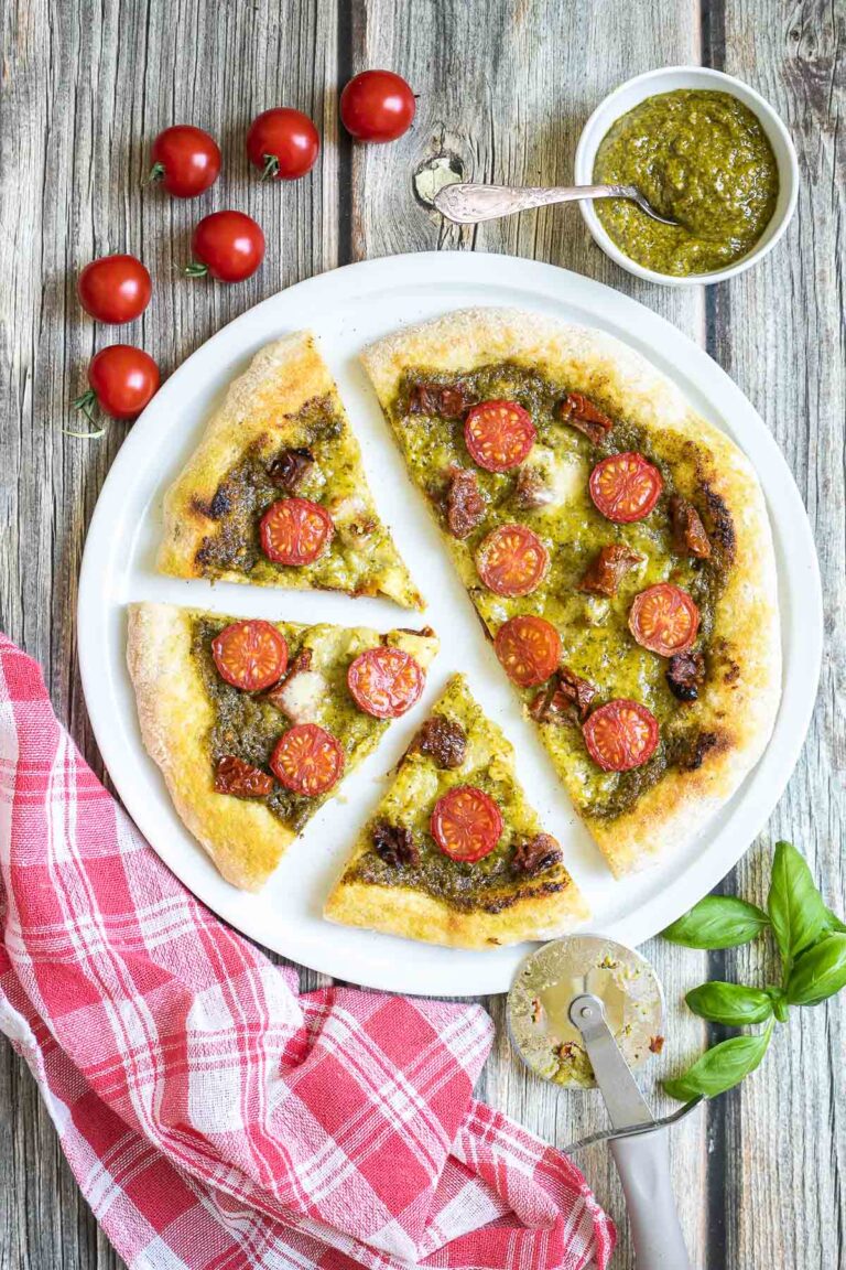 Sliced pizza on a white pizza plate topped with green pesto, melted cheese, sun-dried tomato pieces, and halved cherry tomatoes. More pesto and fresh ingredients next to the plate.