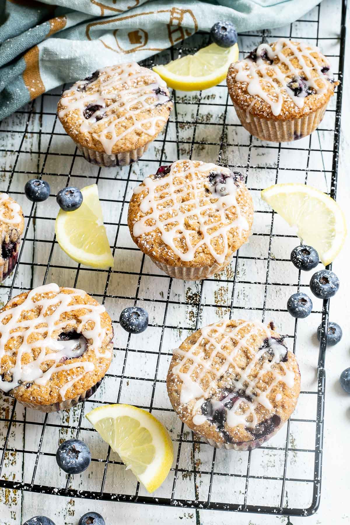 Several lemon muffins with blueberries on a black wire rack drizzled with a white glaze. Lemon slices and fresh blueberries are scattered around them.
