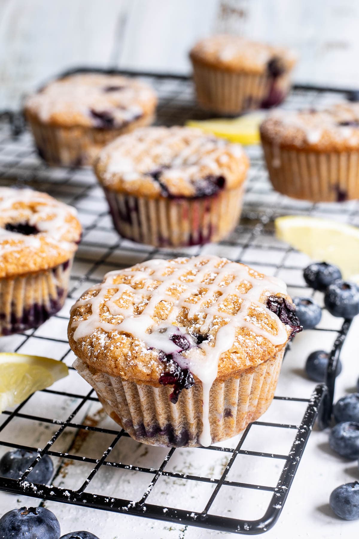 Several lemon muffins with blueberries on a black wire rack drizzled with a white glaze. Lemon slices and fresh blueberries are scattered around them.