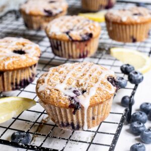Several lemon muffins with blueberries on a black wire rack drizzled with a white glaze. Lemon slices and fresh blueberries are scattered around them.