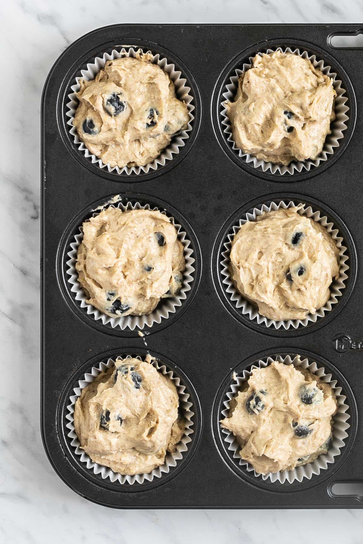 A muffin tin lined with white cupcake liners and filled with light brown batter mixed with fresh blueberries.