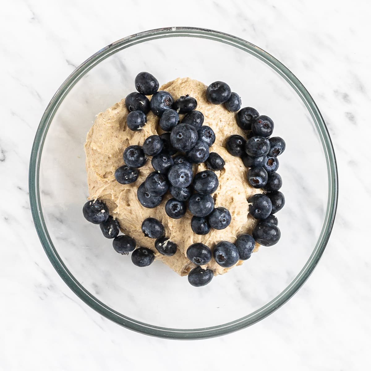 Glass bowl with light brown creamy batter topped with lots of fresh blueberries.