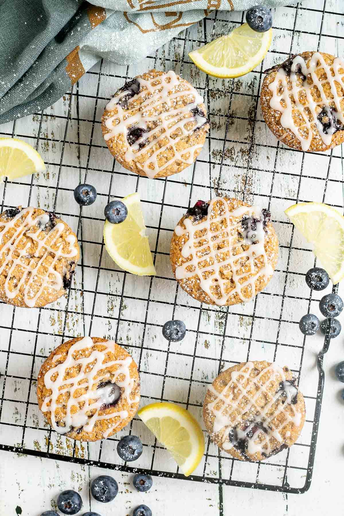 Several lemon muffins with blueberries on a black wire rack drizzled with a white glaze. Lemon slices and fresh blueberries are scattered around them.