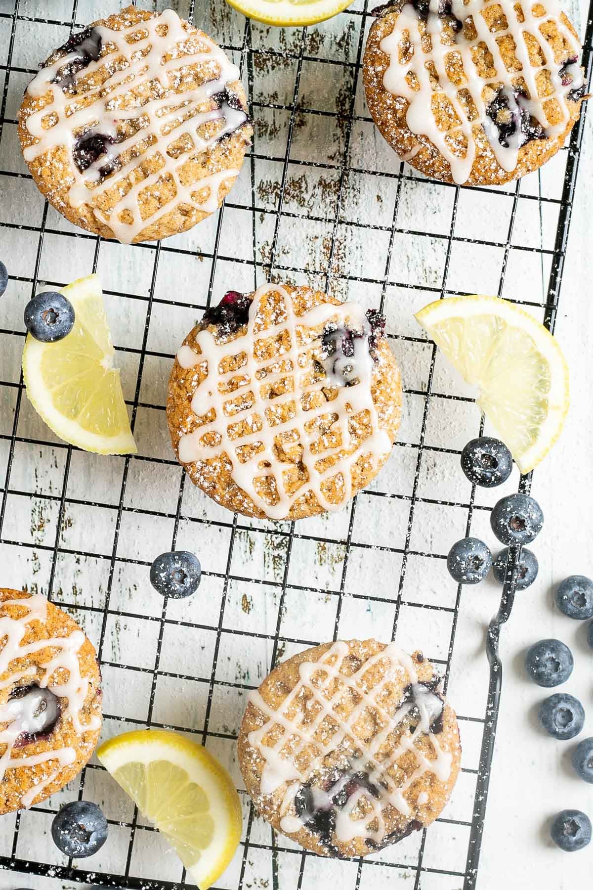 Several lemon muffins with blueberries on a black wire rack drizzled with a white glaze. Lemon slices and fresh blueberries are scattered around them.