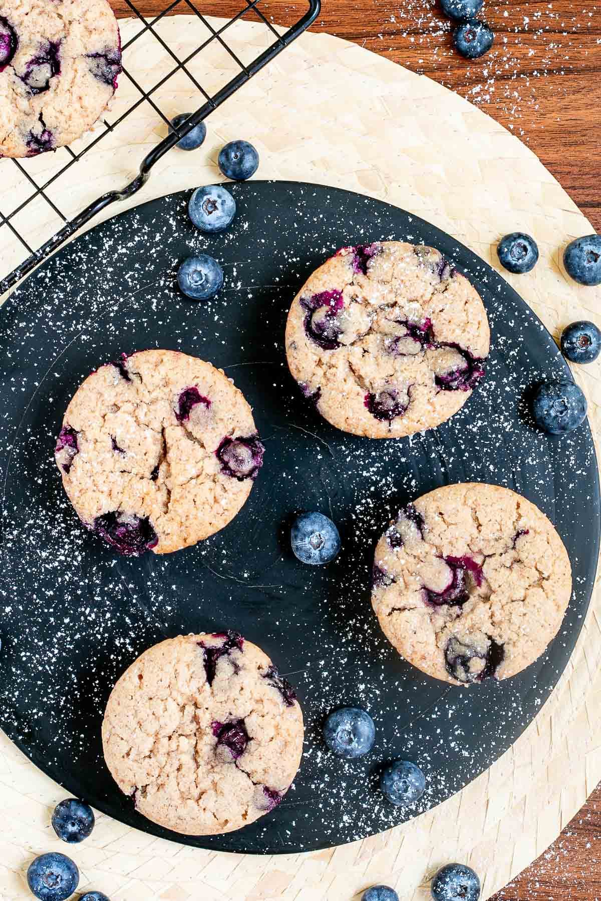 Blueberry muffins on a black board dusted with powdered sugar and surrounded by fresh bluberries.