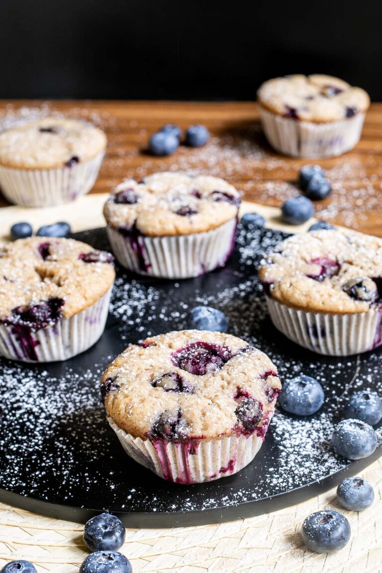 Blueberry muffins on a black board dusted with powdered sugar and surrounded by fresh bluberries.
