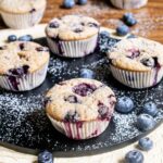 Blueberry muffins on a black board dusted with powdered sugar and surrounded by fresh bluberries.