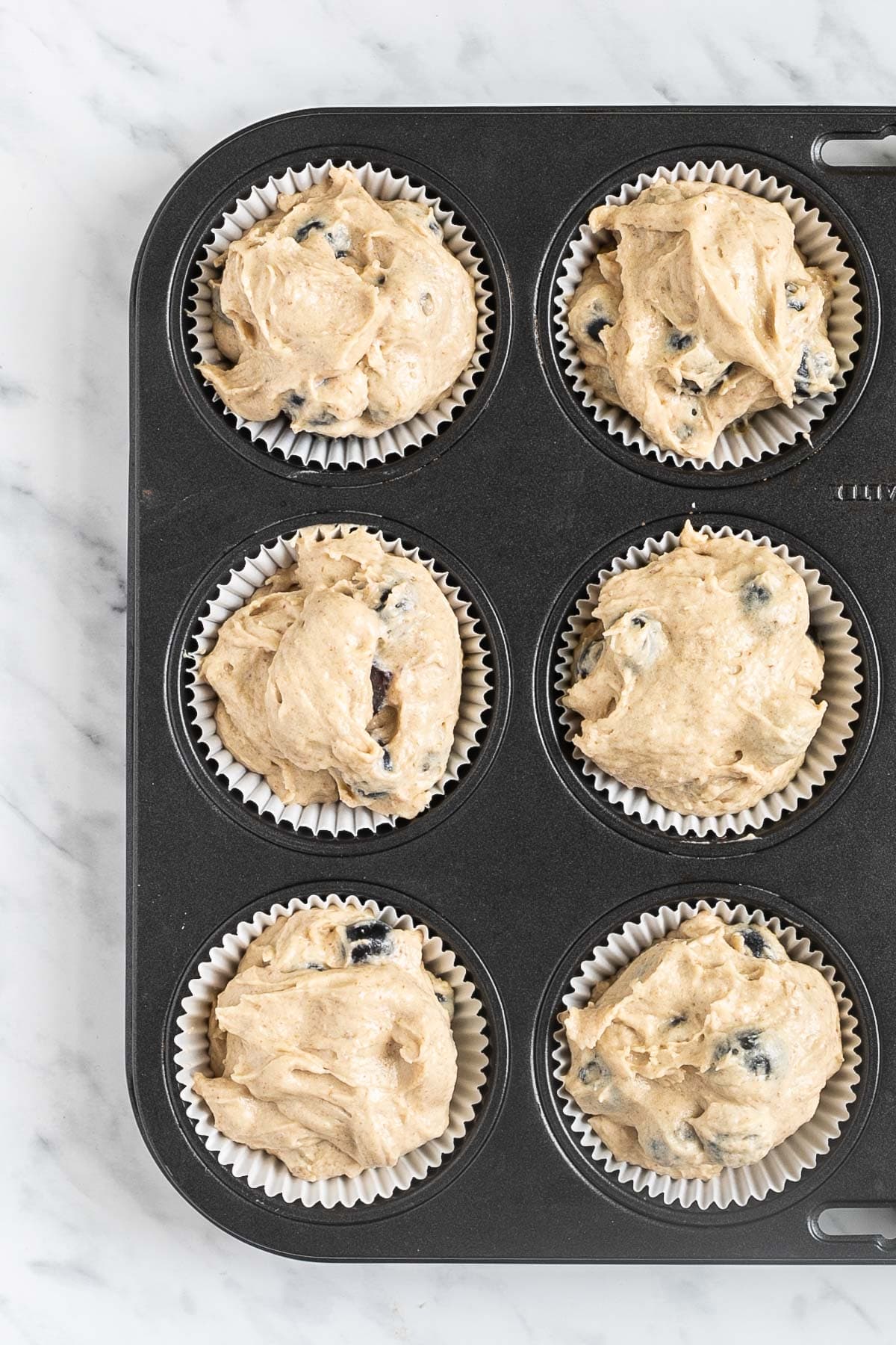 A muffin tin lined with white cupcake liners and filled with light brown batter with blueberries to the top.