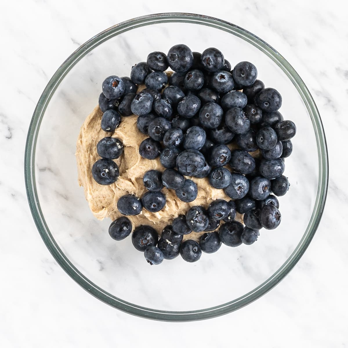 Glass bowl with light brown creamy batter topped with lots of fresh blueberries.