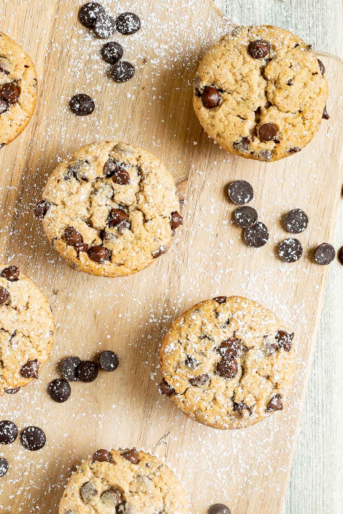 Chocolate chip muffins on a wooden board surrounded by lots of chocolate chips.