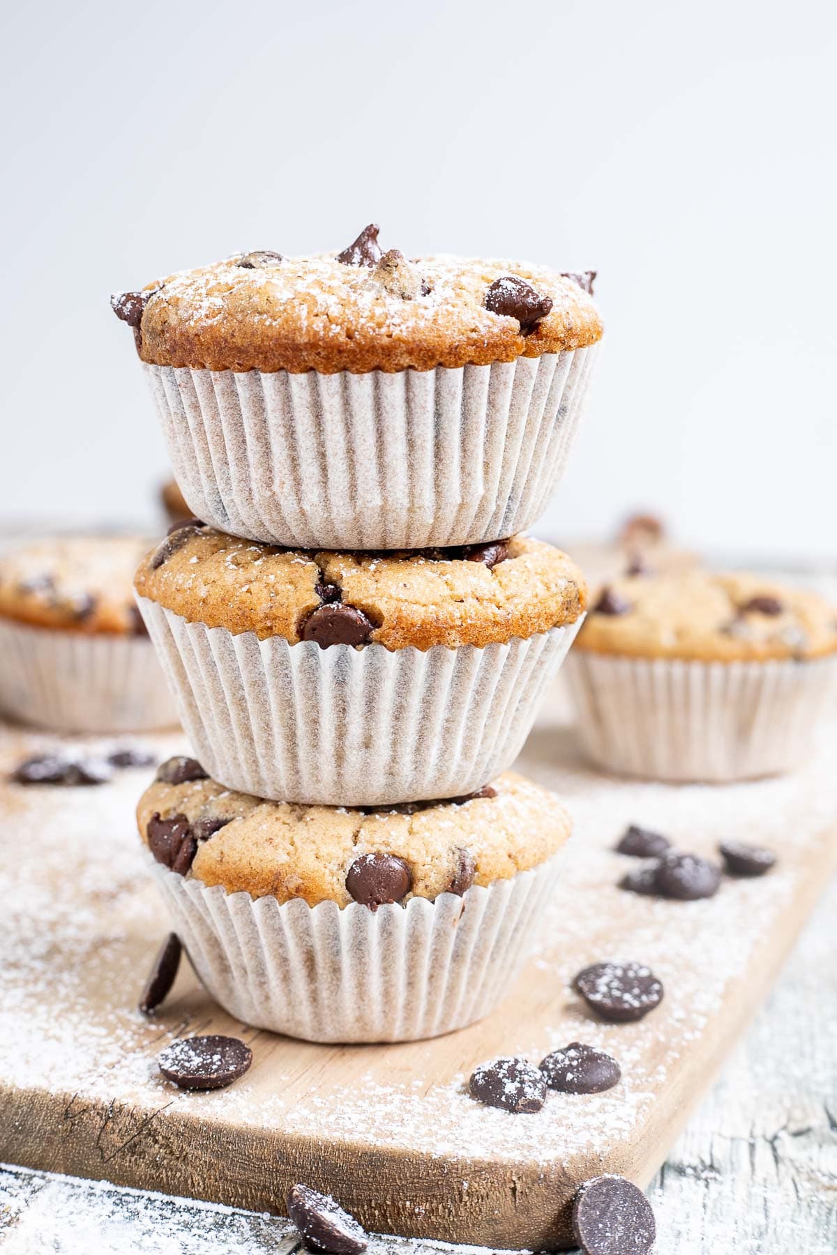 A stack of 3 chocolate chip muffins in paper liners surrounded by other muffins and more chocolate chips.