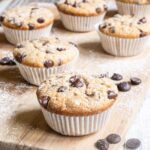 Lightly dusted chocolate chip muffins on a wooden board surrounded by lots of chocolate chips.