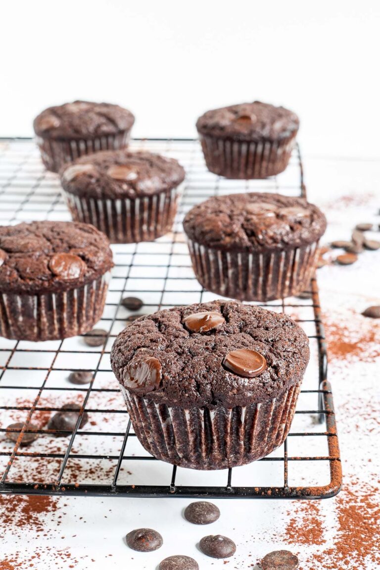 Several chocolate muffins with chocolate chips on a black wire rack. Even more chocolate chips are scattered around and under the rack.