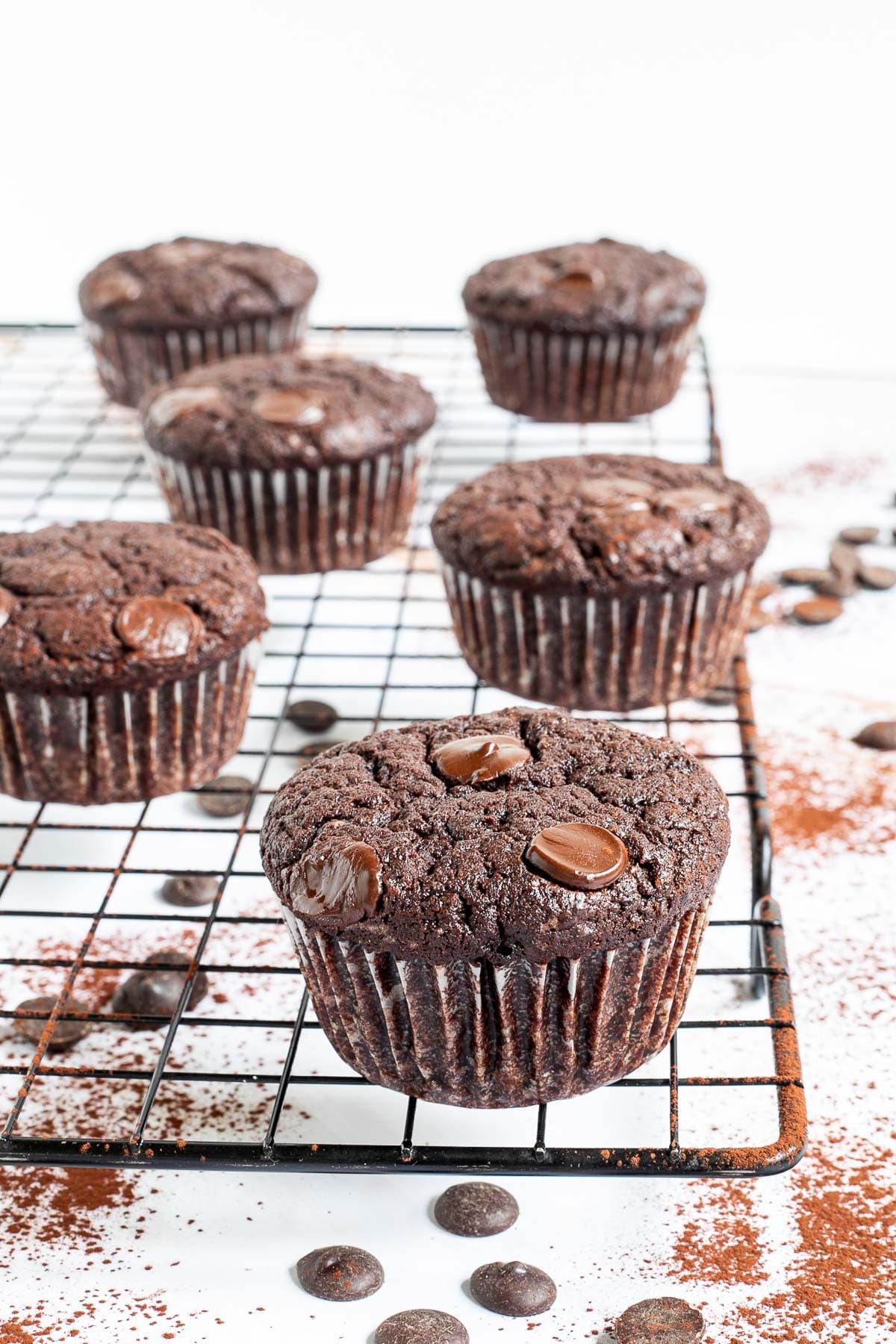 Several chocolate muffins with chocolate chips on a black wire rack. Even more chocolate chips are scattered around and under the rack.