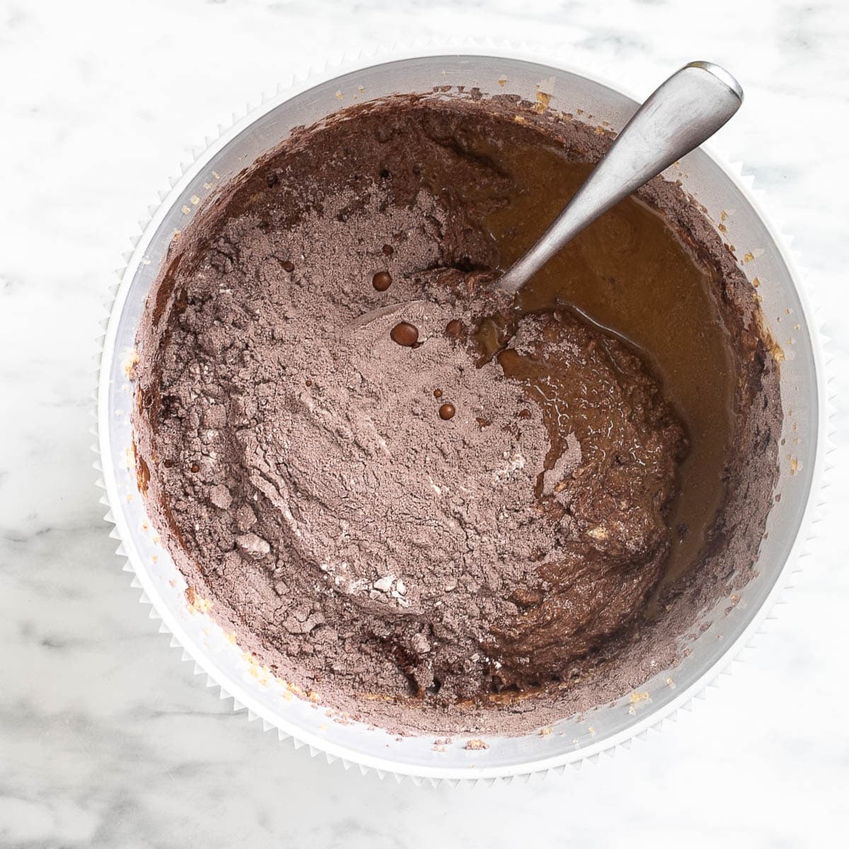 Liquid and a heap of brown cocoa flour mix are on top of a soft dark brown batter in a white bowl.
