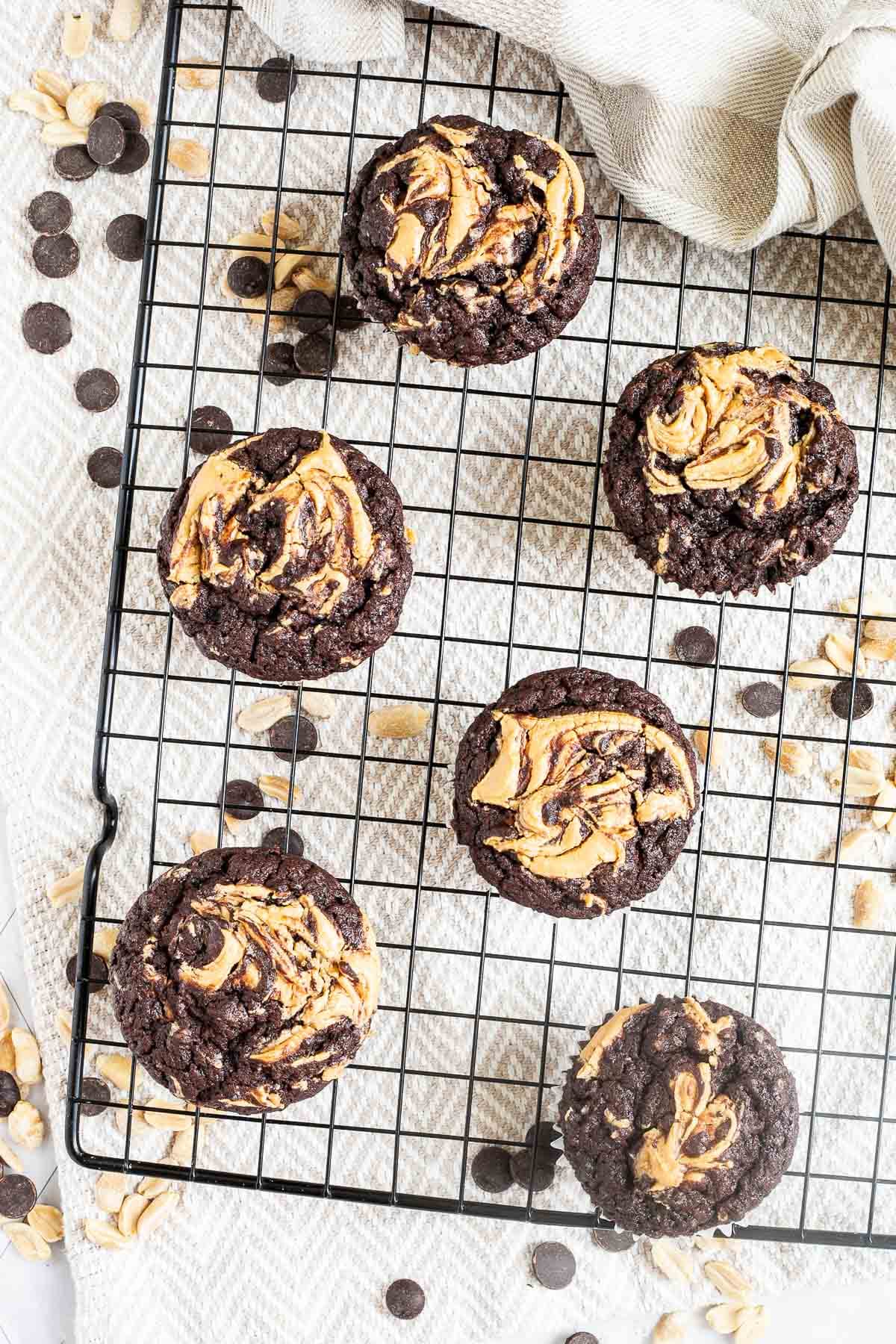 Several chocolate muffins with a peanut butter swirl on top on a black wire rack. Even more chocolate chips and peanuts are scattered around and under the rack.
