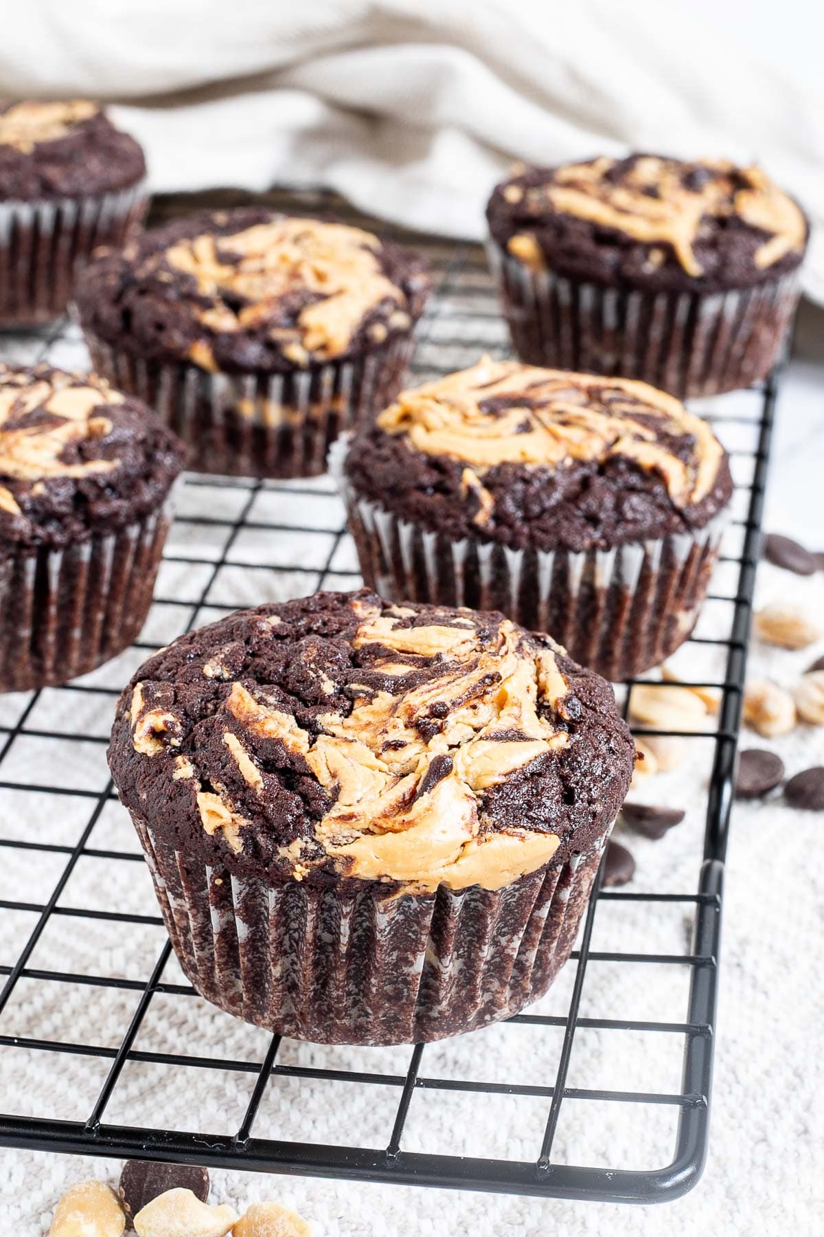Several chocolate muffins with a peanut butter swirl on top on a black wire rack. Even more chocolate chips and peanuts are scattered around and under the rack.