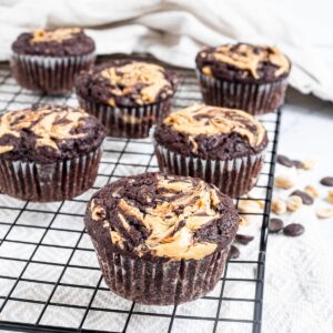 Several chocolate muffins with a peanut butter swirl on top on a black wire rack. Even more chocolate chips and peanuts are scattered around and under the rack.
