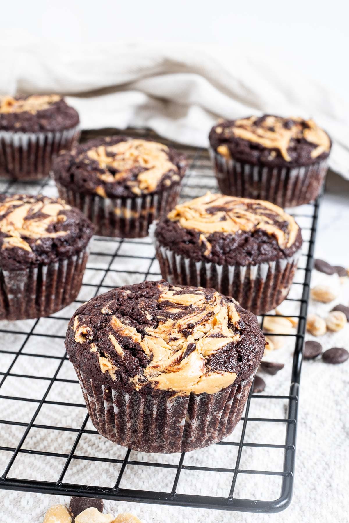 Several chocolate muffins with a peanut butter swirl on top on a black wire rack. Even more chocolate chips and peanuts are scattered around and under the rack.