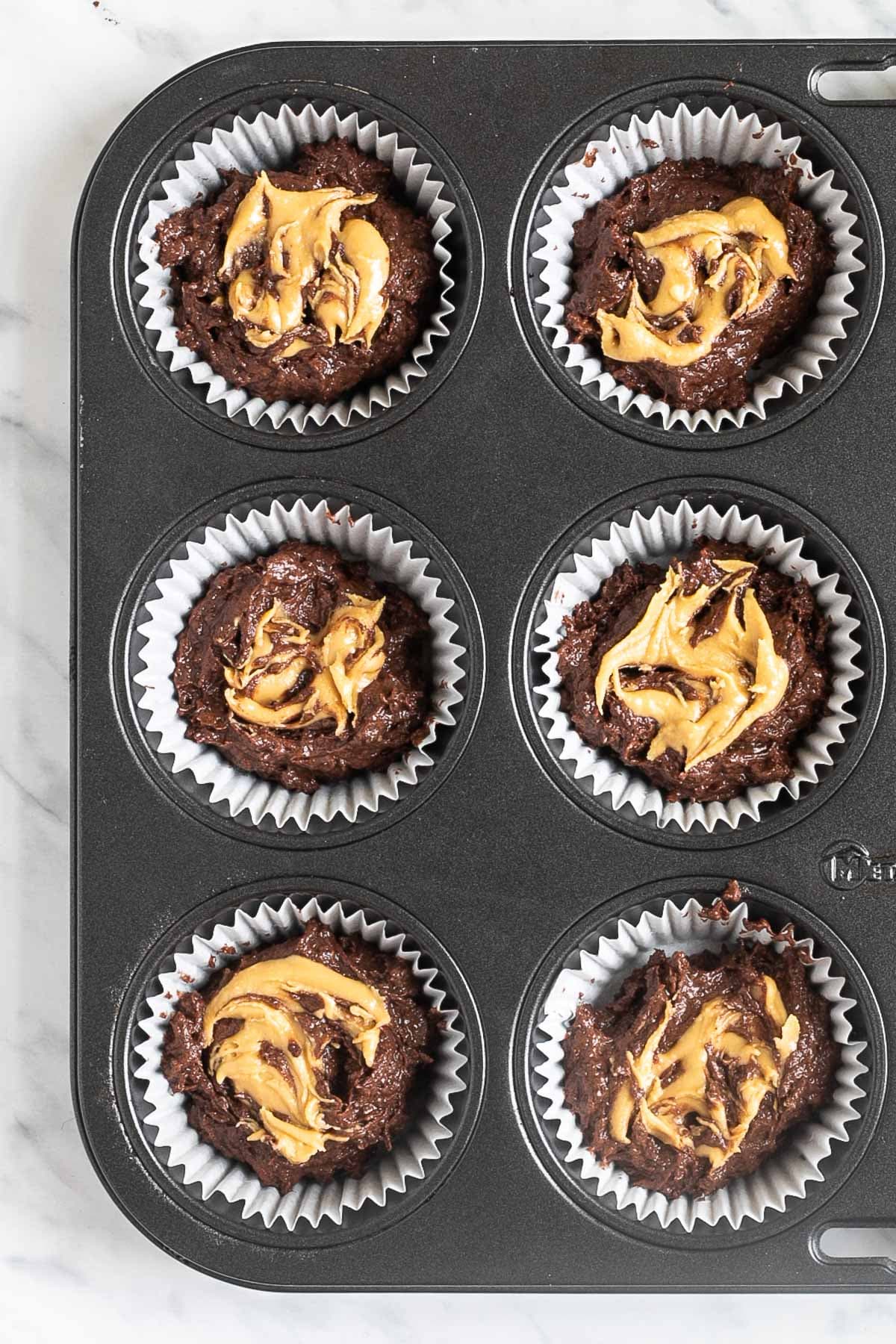 A muffin tin lined with white cupcake liners and filled with dark brown batter topped with a small heap of peanut butter.