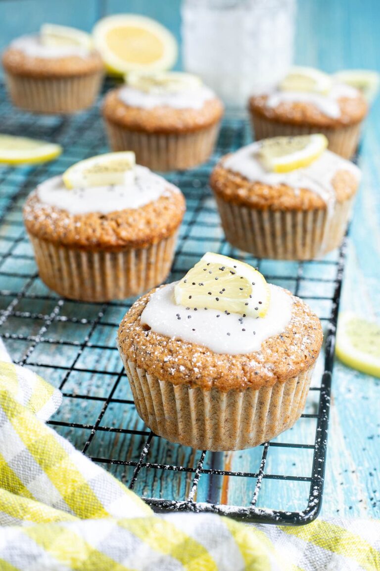 Several lemon poppy seed muffins on a black wire rack topped with a white glaze and a small lemon slices, and sprinkled with poppy seeds. Lemon slices are scattered around them.