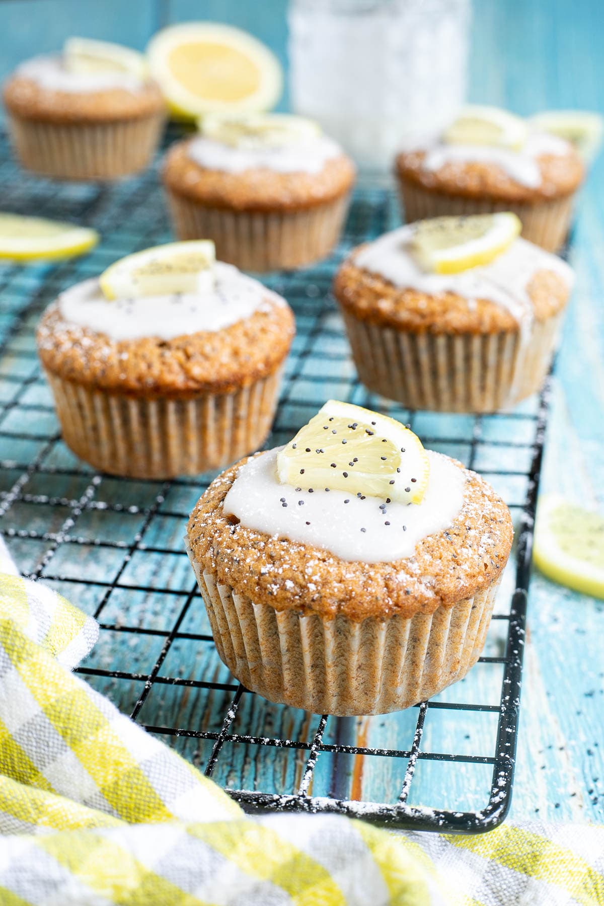 Several lemon poppy seed muffins on a black wire rack topped with a white glaze and a small lemon slices, and sprinkled with poppy seeds. Lemon slices are scattered around them.