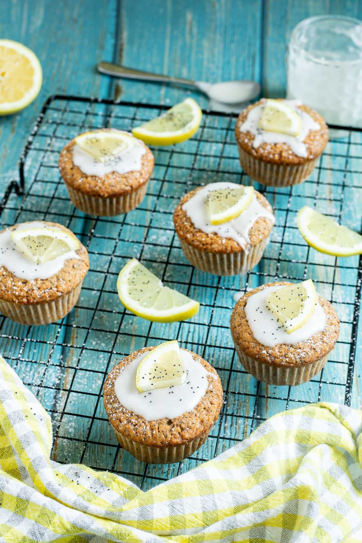Several lemon poppy seed muffins on a black wire rack topped with a white glaze and a small lemon slices, and sprinkled with poppy seeds. Lemon slices are scattered around them.
