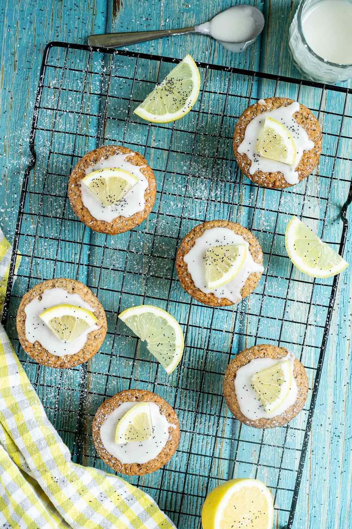 Several lemon poppy seed muffins on a black wire rack topped with a white glaze and a small lemon slices, and sprinkled with poppy seeds. Lemon slices are scattered around them.