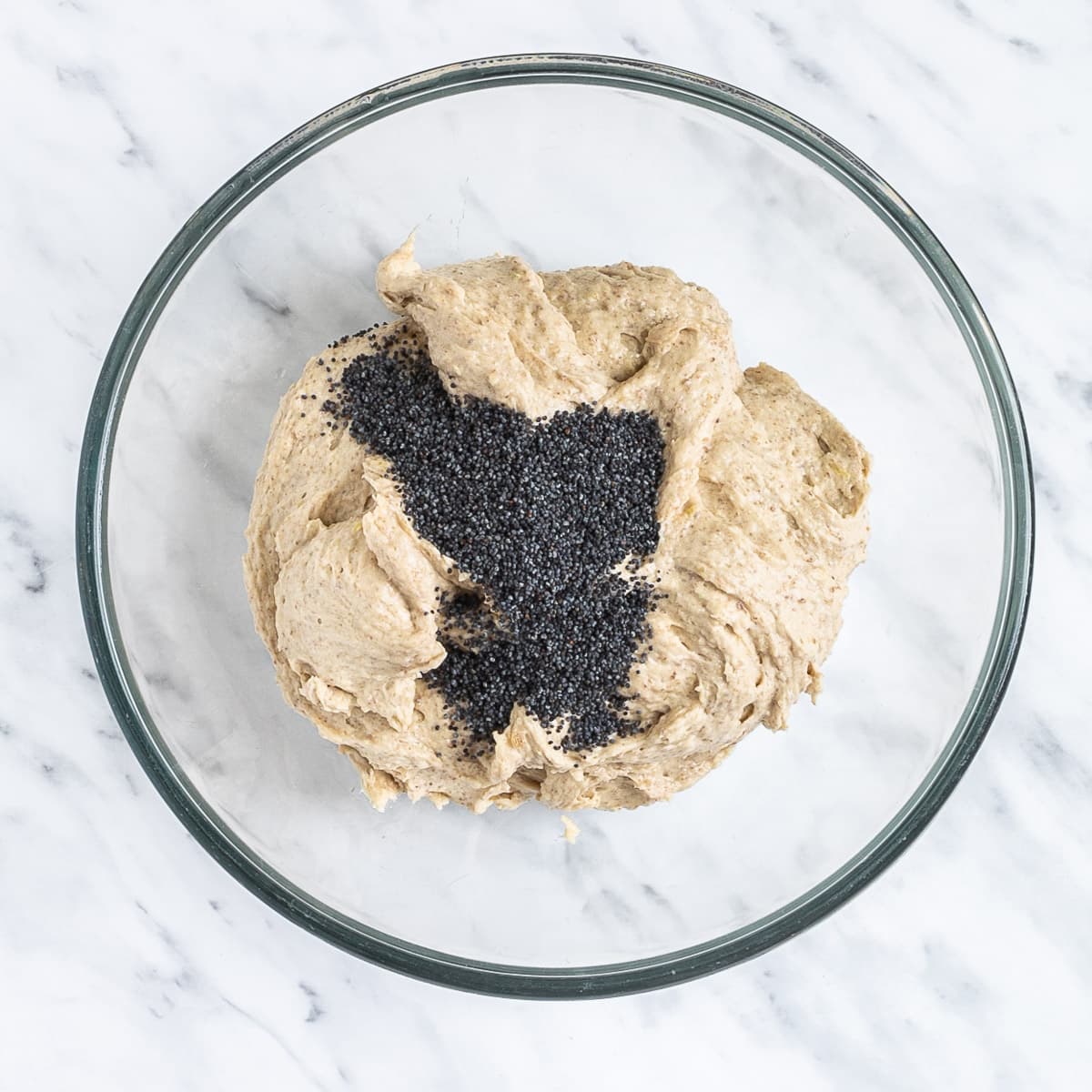 Glass bowl with light brown creamy batter topped with lots of poppy seeds.