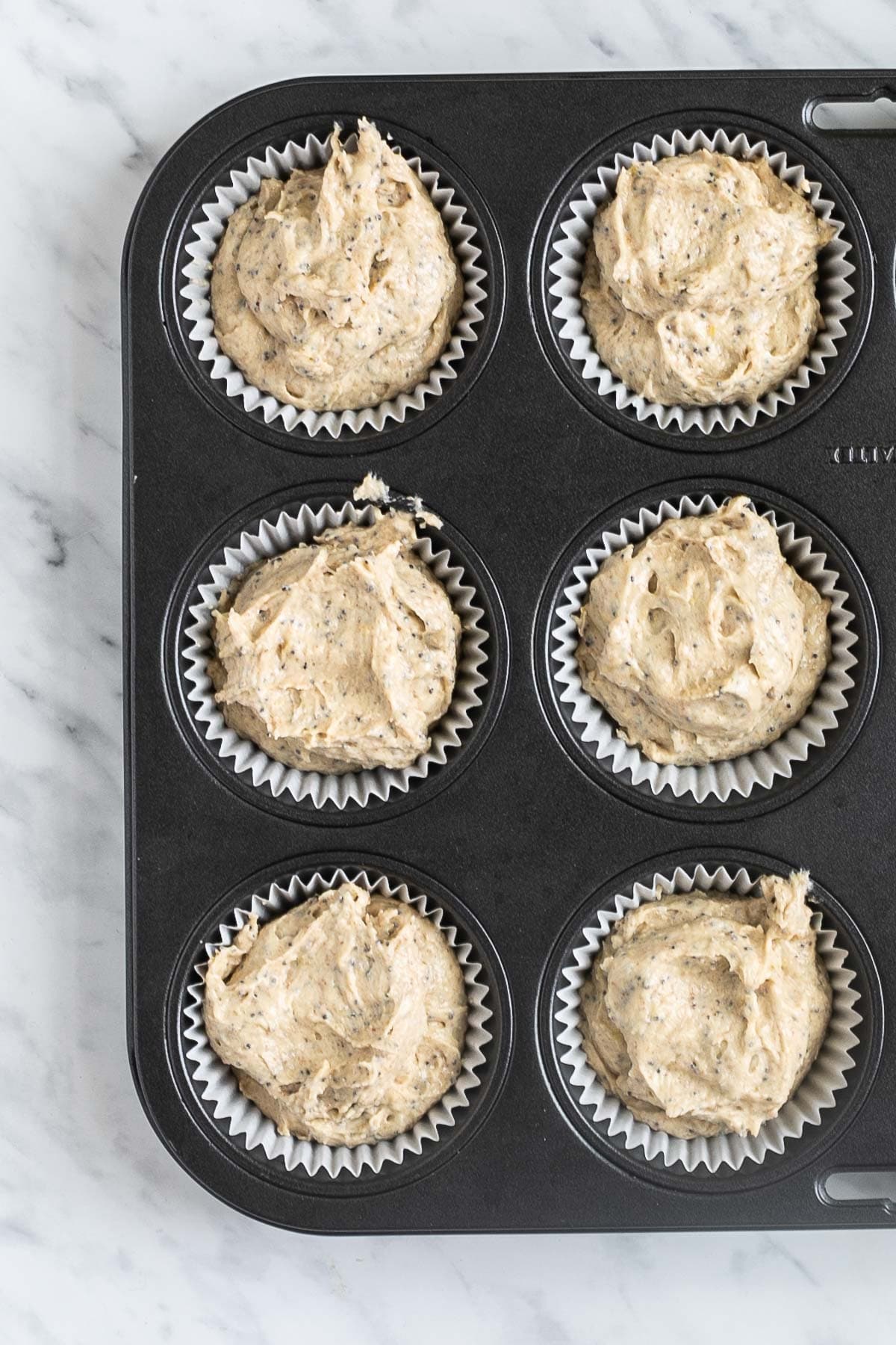A muffin tin lined with white cupcake liners and filled with light brown batter mixed with poppy seeds.
